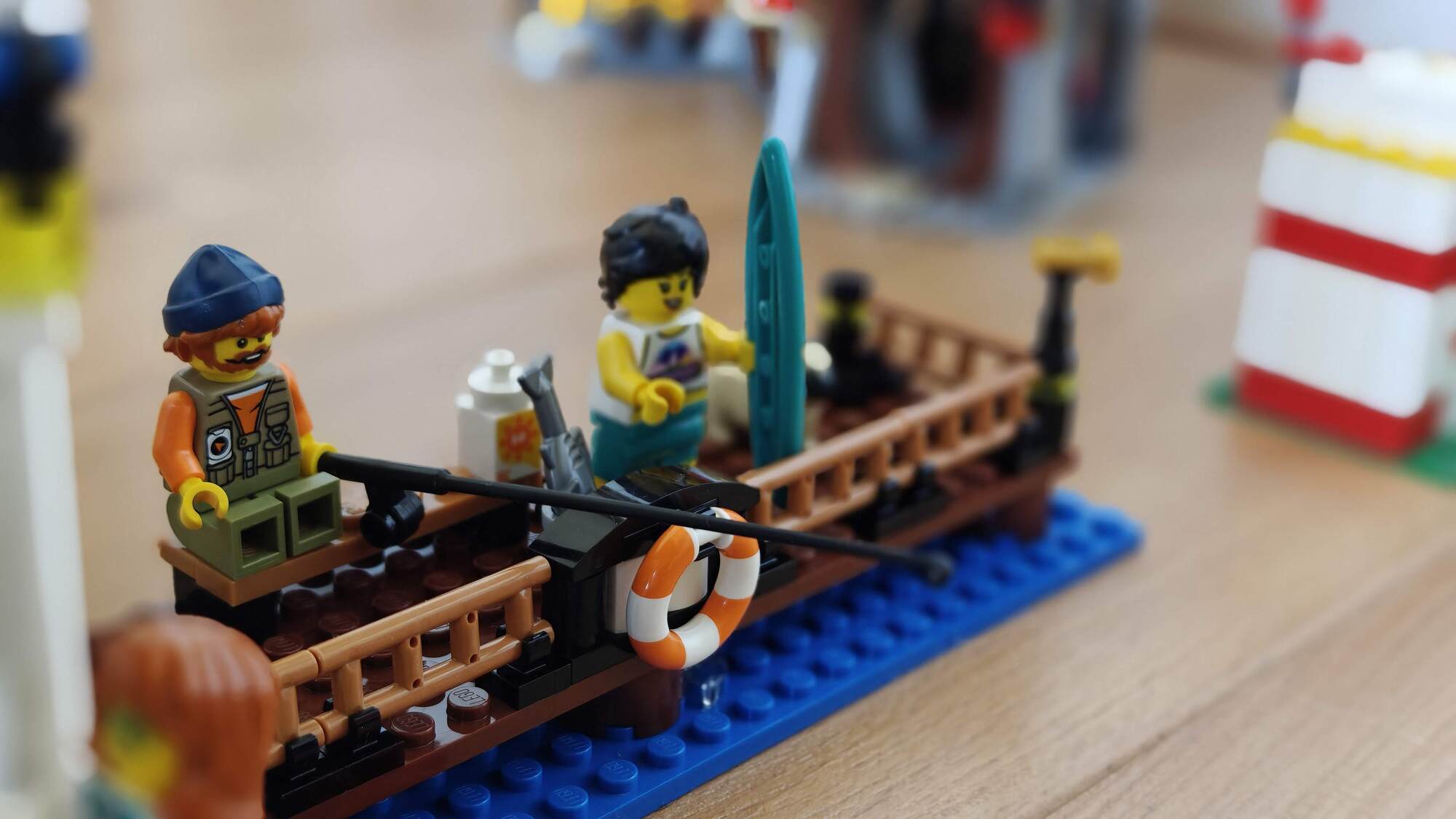 A minifigure sits on the beach with fishing rods, next to another minifigure holding a surfboard