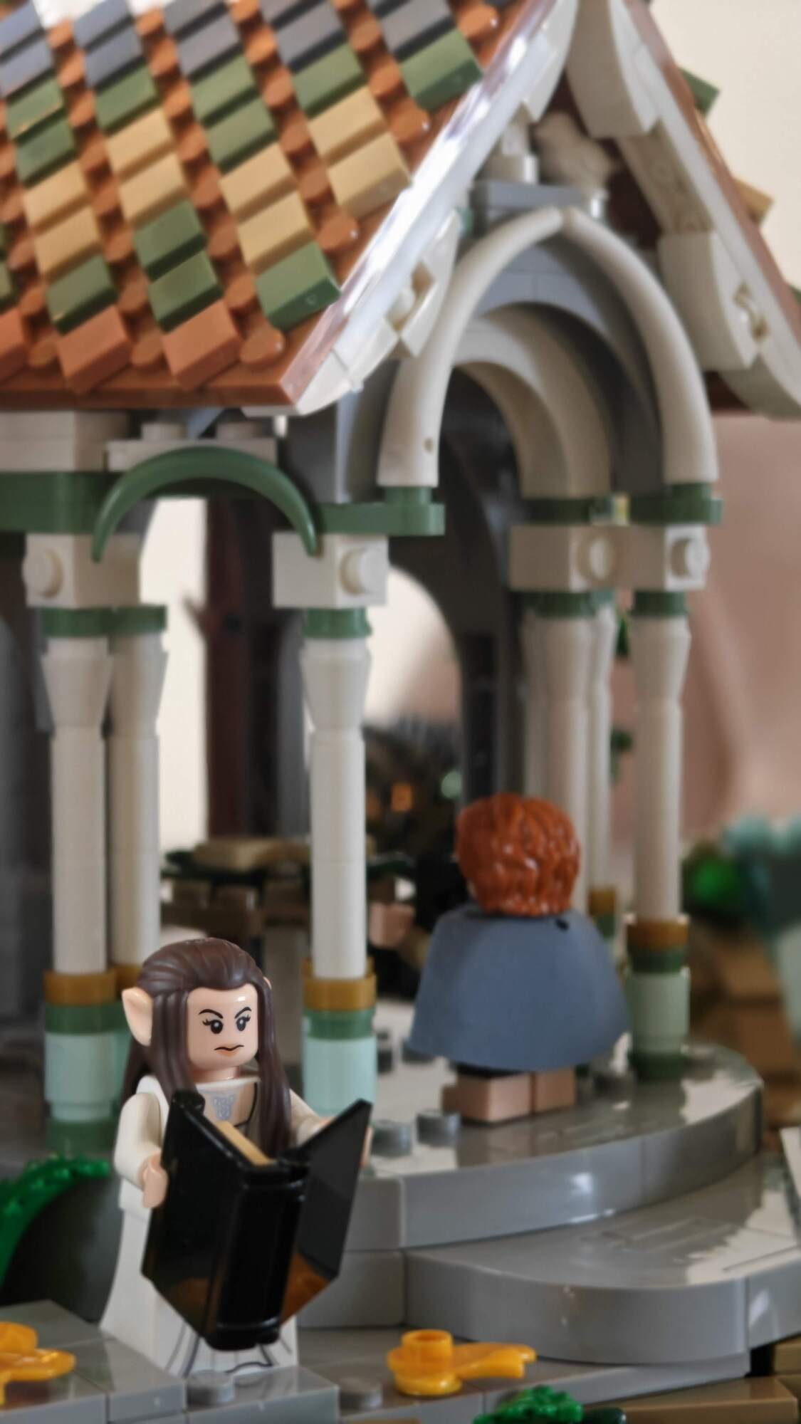Minifig Arwen com um livro na mão está em frente à pequena casa em Rivendell, com Samwise ao fundo